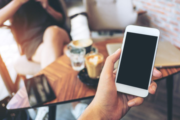 mockup image of hand holding white mobile phone with blank black screen in vintage cafe and blur asi