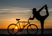 Silhouette Of Girl With Bicycle At Sunset Time. Young Woman Is Doing Yoga Near The Ocean With Bicycle