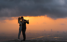 Two Lovers Dancing/embracing On Top Of A Skyscraper Overlooking The City At Sunset. Romantic Setting.