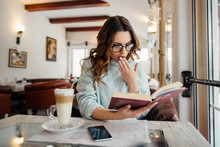 Emotional Girl In Glasses Reading Book In Cafe While Drinking Coffee