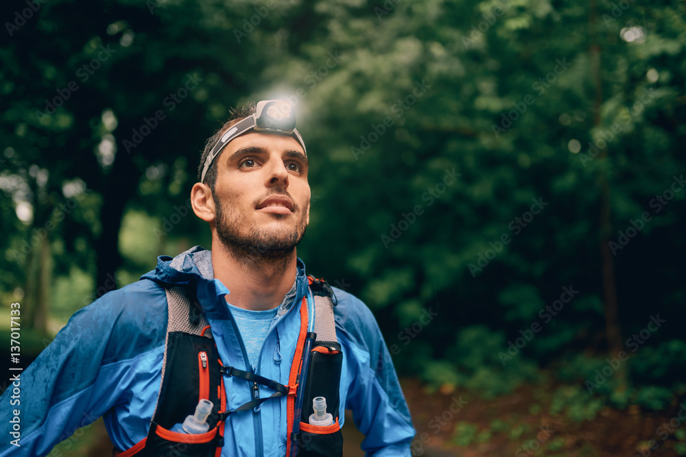 Fit male jogger with a headlamp rests during training for cross country trail race in nature park. - obrazy, fototapety, plakaty 
