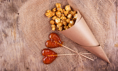 Two lollipop in the shape of a heart and a paper bag of caramel popcorn on wooden background. Fast food and unhealthy food.