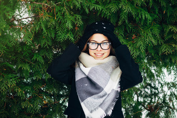 Wall Mural - Portrait of a beautiful girl with blue eyes in black glasses in a hat and coat with scarf in the winter park