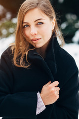 Wall Mural - Portrait of a beautiful girl with blue eyes in a black coat with in the winter park on the background of christmas tree