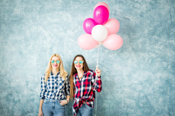 Two female friends in checkered shirts having fun with air balloons on the blue wall background