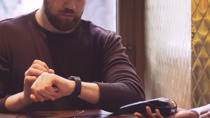 Poster - Man in sweater sitting by the table, looking at check, using wristwatch and paid the bill by means of wireless payment technology in cafe