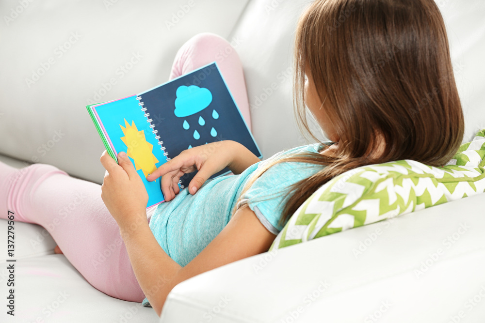 Attractive little girl with book sitting on sofa at home