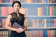 © nakophotography - Well dressed and cared for African American woman looking at camera while holding her notebook and digital tablet in her hand in front of her with a shelf of books behind her in the background.
