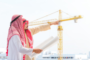 Portrait of arabian engineer talking about saudi job and verify the blueprint with commitment to success at construction site with crane background