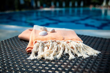 The Concept Of Summer Accessories Close-up Of A White And Orange Turkish Peshtemal / Towel And White Seashells On Rattan Lounger With Blue A Swimming Pool As Background.