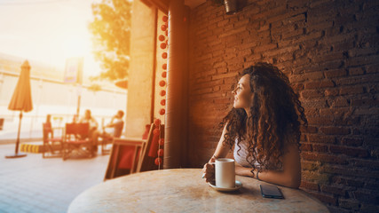 Young pretty curly brunette woman drinking cappuccino while relaxing in cozy cafe, pensive female enjoying cup of tea while sitting in coffee shop during her recreation time with smartphone near her