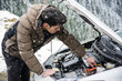 © theartofphoto - Young man near car with open hood inspecting engine in winter. Snowy forest on background