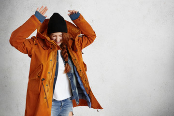 Fashionable young woman wearing red winter coat, black hat and ragged jeans raising hands in air while having fun indoors against grey studio wall background with copy space for your content