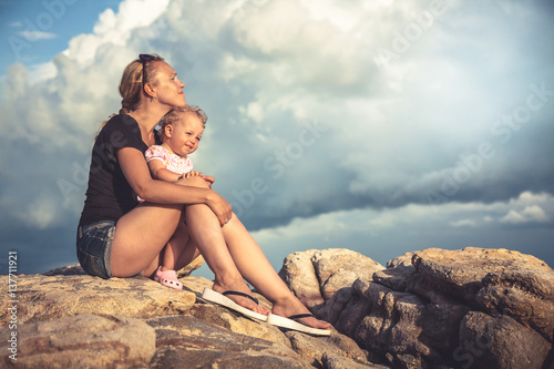 Fotografie, Obraz Loving young mother embrace her baby sitting on rocks with dramatic sky and clou