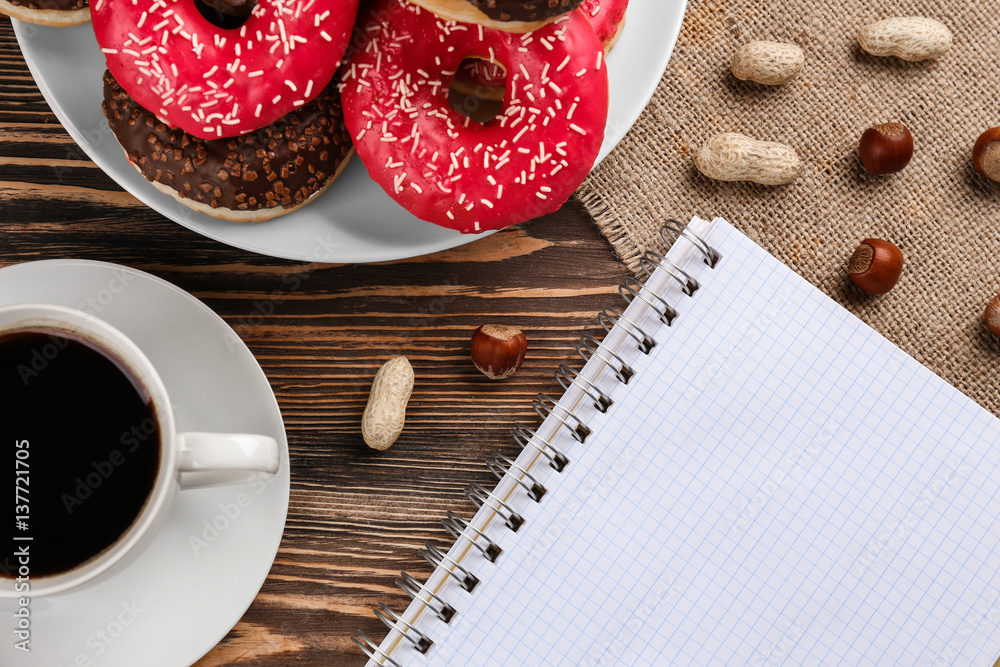 Tasty donuts with cup of coffee and notebook on wooden table