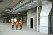 © pressmaster - Group of three workmen wearing protective helmets and vests on construction site: walking among concrete walls at basement floor of unfinished building with foreman inspector