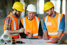 Team Of Construction Workers Wearing Protective Helmets And Vests Discussing Project Details With Executive Supervisor Standing At Table With Blueprints