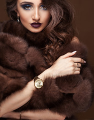 Luxury elegant woman in fur coat, golden earrings and watches. Glamour beautiful look. Rich lady over dark chocolat background.