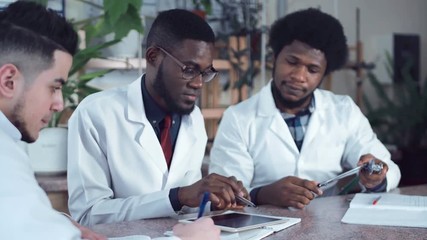 Young African American men doctors in white coats sitting at table in hospital and working with tablet and stethoscope