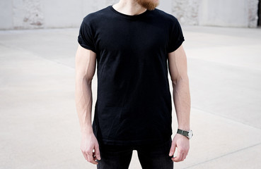 Young muscular bearded man wearing black tshirt and jeans posing in center of modern city. Empty concrete wall on the background. Hotizontal mockup, front view