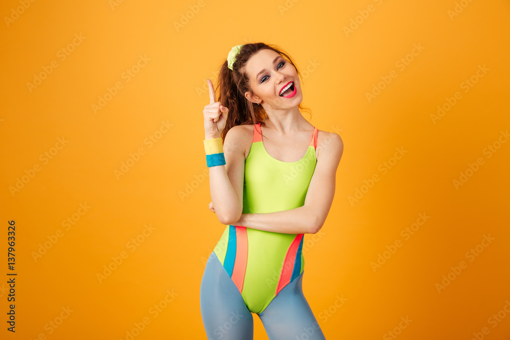 Smiling cute young woman athlete standing and pointing up - obrazy, fototapety, plakaty 