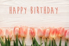 Happy Birthday Text Sign On Pink Tulips On White Rustic Wooden Background Flat Lay. Spring Top View Of Flowers In Soft Morning Light With Space For Text. Greeting Card Concept