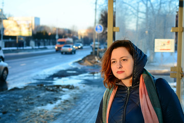 Young woman on a bus stop, caucasian girl with red hair waiting for the city bus, european female wearing hooded coat in the city bus station, sunset in the city