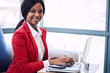 © nakophotography - Black business woman looking at camera while smiling, with her hands still on the keyboard of her laptop computer where she was sitting working before looking up at camera.