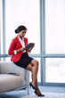 © nakophotography - Young black businesswoman sitting on the edge of a sofa in a business lounge while looking at the screen of the digital tablet that she is holding in her hands.