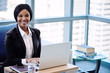 © nakophotography - African business woman smiling at camera while she was busy working on her laptop in the business lounge of her modern co working office, with large windows and the cityscape in the background.