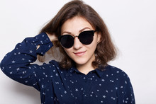 People And Lifestyle Concept. Portrait Of Fashionable Woman Wearing Round Hipster Sunglasses And Stylish Shirt Looking At The Camera, Smiling, Holding Her Hand On Hair. Cute Female Model.