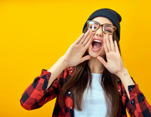 Hipster Girl Screaming Like In Megaphone Holding Hands Near Her Face With Open Mouth. Young Woman Touts Everyone Over Yellow Background.