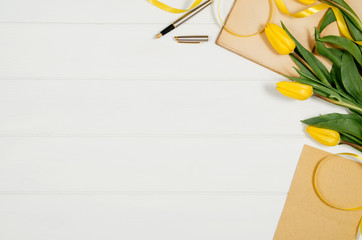 Poster - Background with yellow tulips, blank notepad on white wooden table top view. Creative woman's workspace concept with copy space. Product photograph taken from above with frame composition