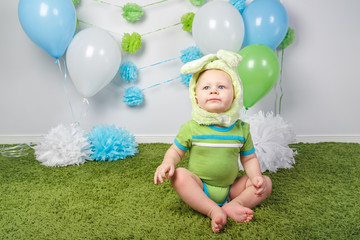 Portrait of cute adorable Caucasian baby boy in holiday Easter bunny rabbit costume with large ears,  dressed in green clothes onesie, sitting on soft fluffy rug carpet in studio on white background.