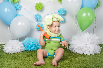 Portrait of cute adorable Caucasian baby boy in holiday Easter bunny rabbit costume with large ears,  dressed in green clothes onesie, sitting on soft fluffy rug carpet in studio, holding red carrot..