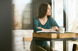 © sata_production - Young woman sits near the window at the cafe and working, she is making notes in her notepad, her laptop is open