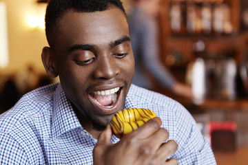 Close-up shot of handsome young dark-skinned man in checkered shirt having tempted look opening his mouth widely, ready to bite doughnut during lunch at restaurant. People, food and leisure concept