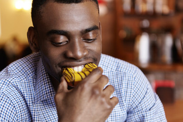 Headshot of attractive young African-American businessman biting doughnut, enjoying junk food during coffee break in blurred restaurant interior. Hungry dark-skinned student eating dessert at cafe