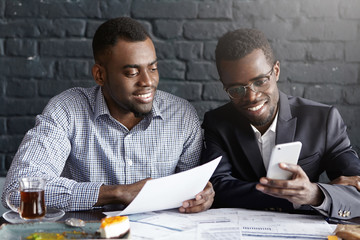 People, cooperation and teamwork concept. Two happy confident dark-skinned colleagues surfing internet on mobile phone while having break during brainstorming session, sharing business ideas