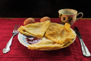 Pancakes with cherry jam, cutlery, eggs and a cup on a red background