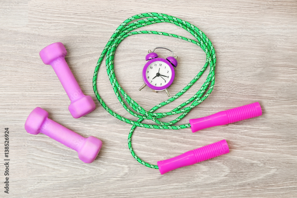Alarm clock with dumbbells and skipping rope on wooden background
