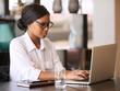 © nakophotography - Accomplished young black businesswoman busy typing on her notebook computer while seated at her dining table at home, weating prescription glasses to see better.