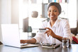 © nakophotography - Good looking young black woman smiling at the camera while seated in front of her computer at her dining room table behind her notebook where she is making online payments with her credit card.