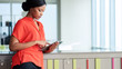 © nakophotography - Attractive businesswoman wearing a bright orange blouse while looking at the screen of the digital electronic tablet that she is busy using with large bright windows in the background.