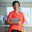 Square portrait of black entrepreneur holding a digital tablet while smiling happily at the camera wearing a bright orange shirt while standing in her usual business environment.