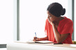 © nakophotography - Self employed black female entrepreneur busy writing in her notebook while seated at a desk with a large bright window behind her in the background.