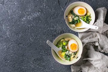 Two bowls with asian style soup with scrambled eggs, half of marinated egg, spring onion, spinach served with white spoons and textile over black texture concrete background. Top view with space