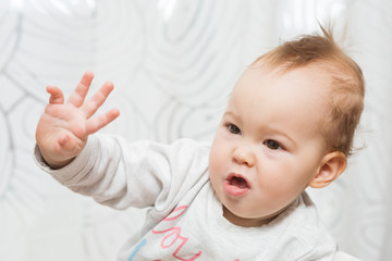 Wall Mural - Eleven months old baby girl sitting in a stool making funny faces into the camera and raising her right arm in the air as if trying to stop someone or reach for something