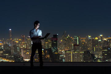 Wall Mural - Asian businessman standing and using the laptop over the cityscape background at night time, Business success and technology concept