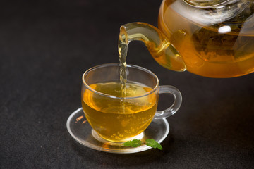 Wall Mural - Pouring tea. Cup of hot tea and tea leaf on black stone background.